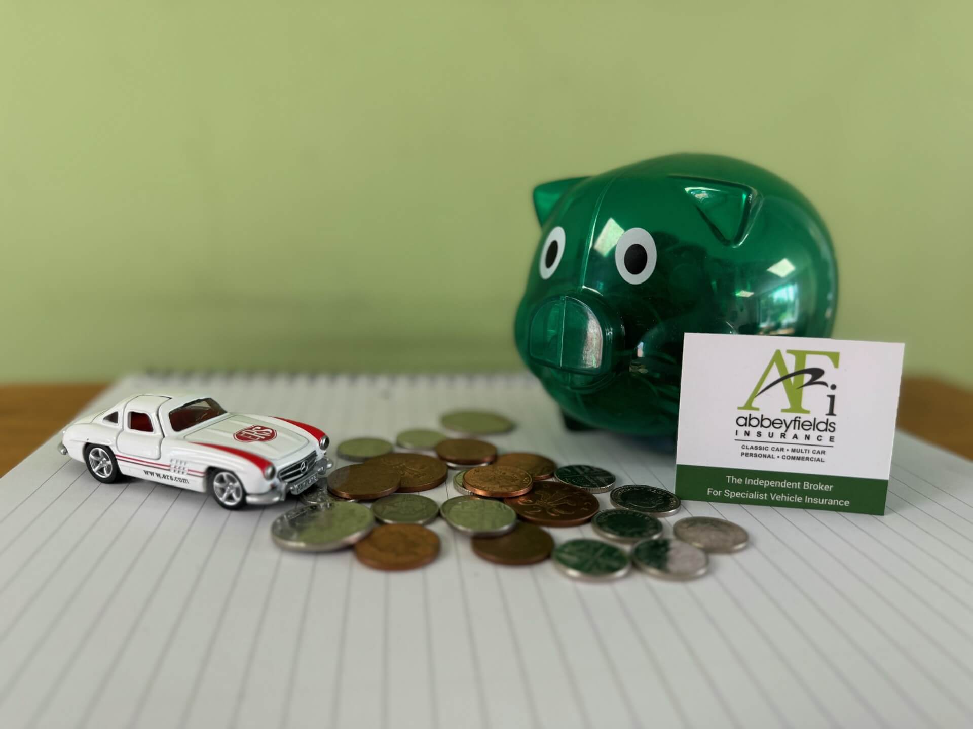 Minature toy car on a notepad next to some coins that have fallen out of a green piggy bank